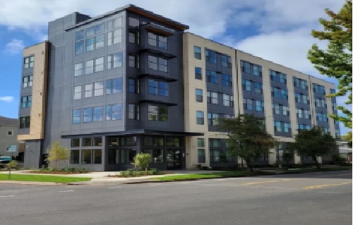 Ferry Street Flats Eugene is a modern, five-story apartment building with large windows and gray and beige exterior panels, located on a corner with trees and a sidewalk in front.