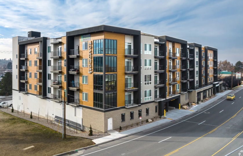 Modern five-story apartment building, The Gardens on 40th Ogden, features balconies, large windows, and a mix of wood and gray exterior panels along a wide street with parked cars and nearby greenery.