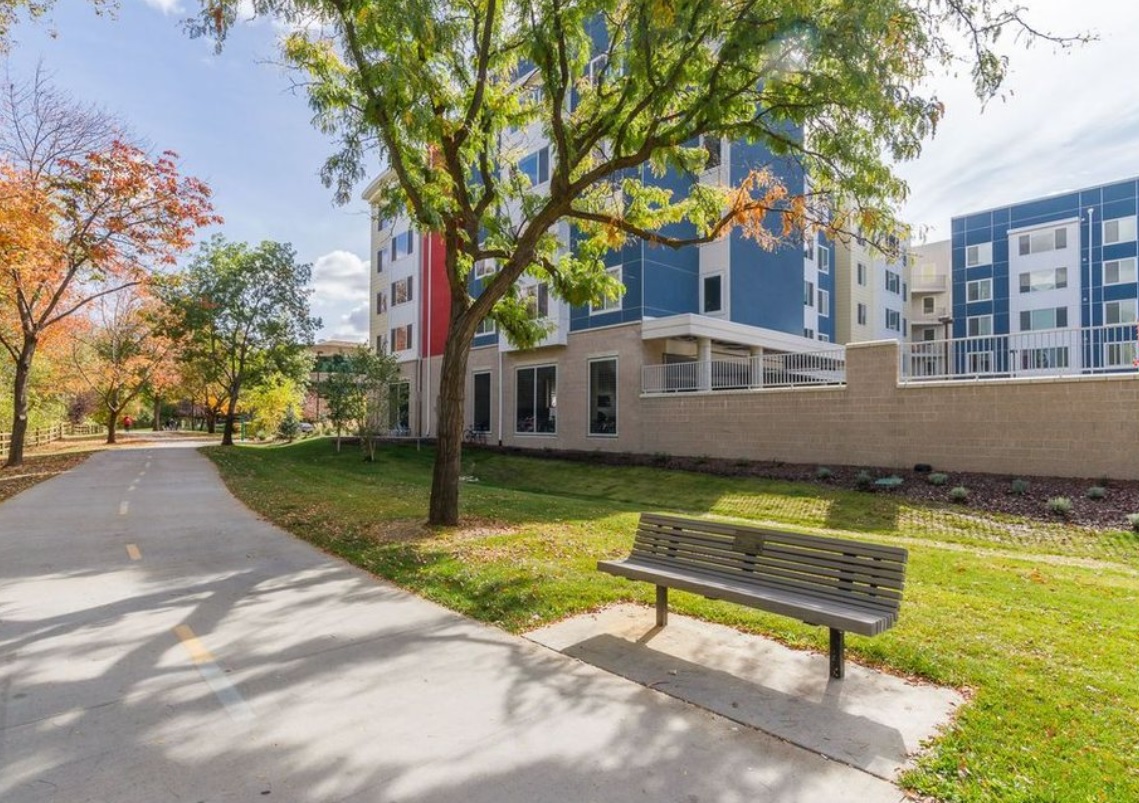 A paved path runs beside Green Leaf River Edge Apartments Boise, a modern building with blue and white panels, bordered by green grass, trees with autumn leaves, and a wooden bench in the foreground.