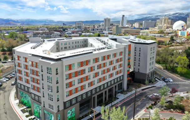 A modern, multi-story apartment building with orange and gray accents stands on a curved street in HERE Reno, city skyline and distant mountains visible in the background under a partly cloudy sky.