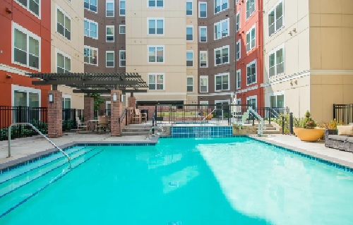 Outdoor swimming pool surrounded by modern Hayward apartments, with poolside seating, tables under a pergola, and potted plants. The bright, inviting area suggests a welcoming community perfect for student accommodation.