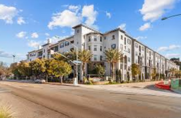 The Iconic on Alvarado is a modern, multi-story apartment building with a white and beige exterior, standing on a sunny street corner surrounded by palm trees and greenery under a blue sky with scattered clouds.