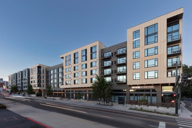 A modern, multi-story apartment building with large windows and balconies sits along a quiet city street at dusk. The ground floor features commercial spaces, and trees line the sidewalk.