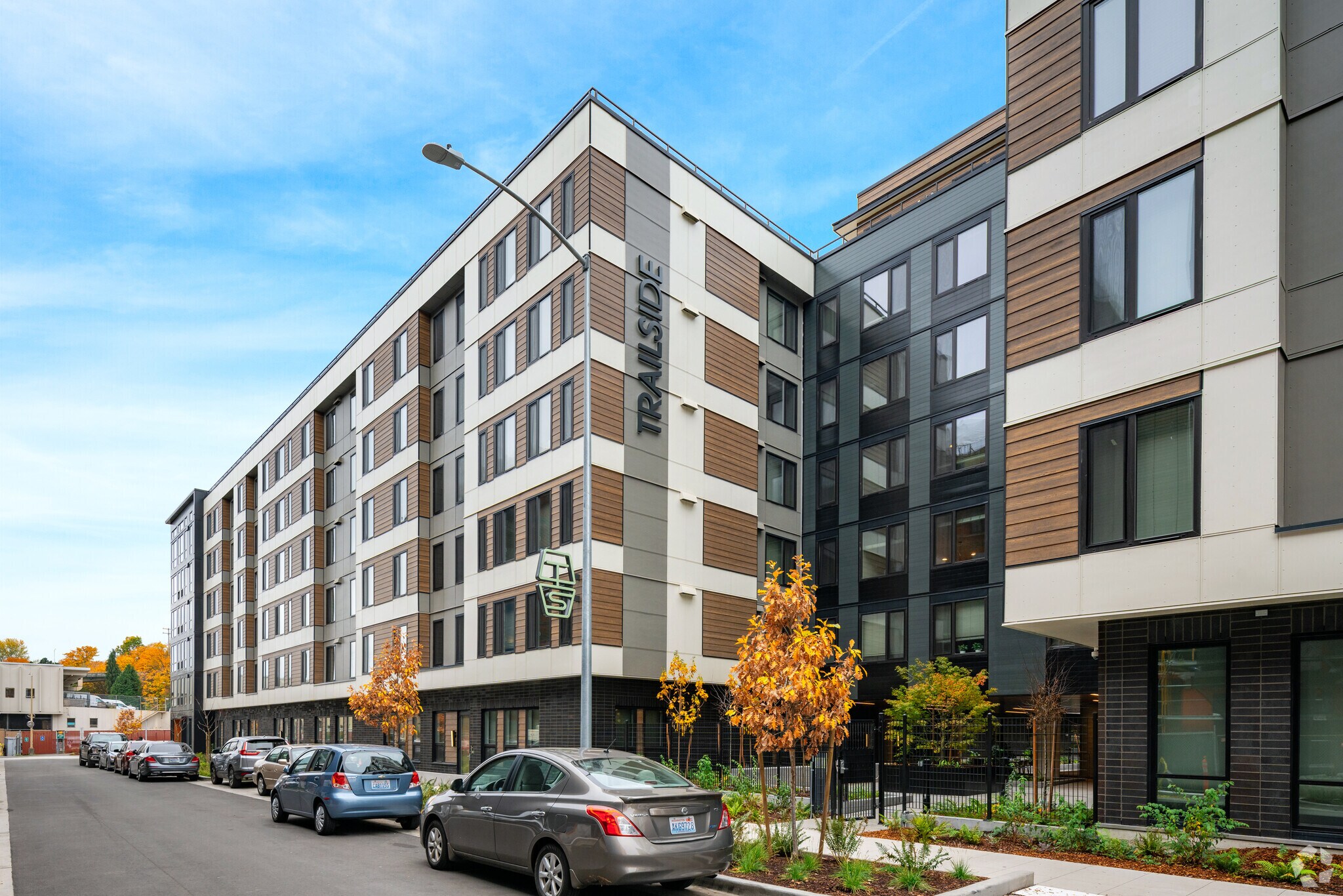 A modern five-story apartment building named Riverside with large windows and brown, white, and gray exterior panels. Parked cars line the street and small trees grow along the sidewalk.