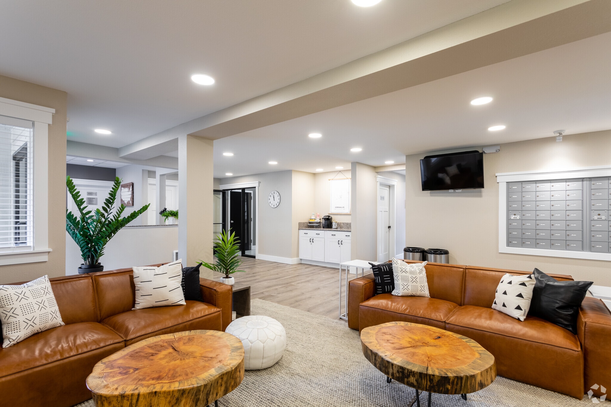 Modern lounge at Oak Vale Apartments with brown leather sofas, wooden coffee tables, patterned pillows, indoor plants, a wall-mounted TV, mailboxes, and a small kitchen area in the background. Warm lighting creates a cozy atmosphere.