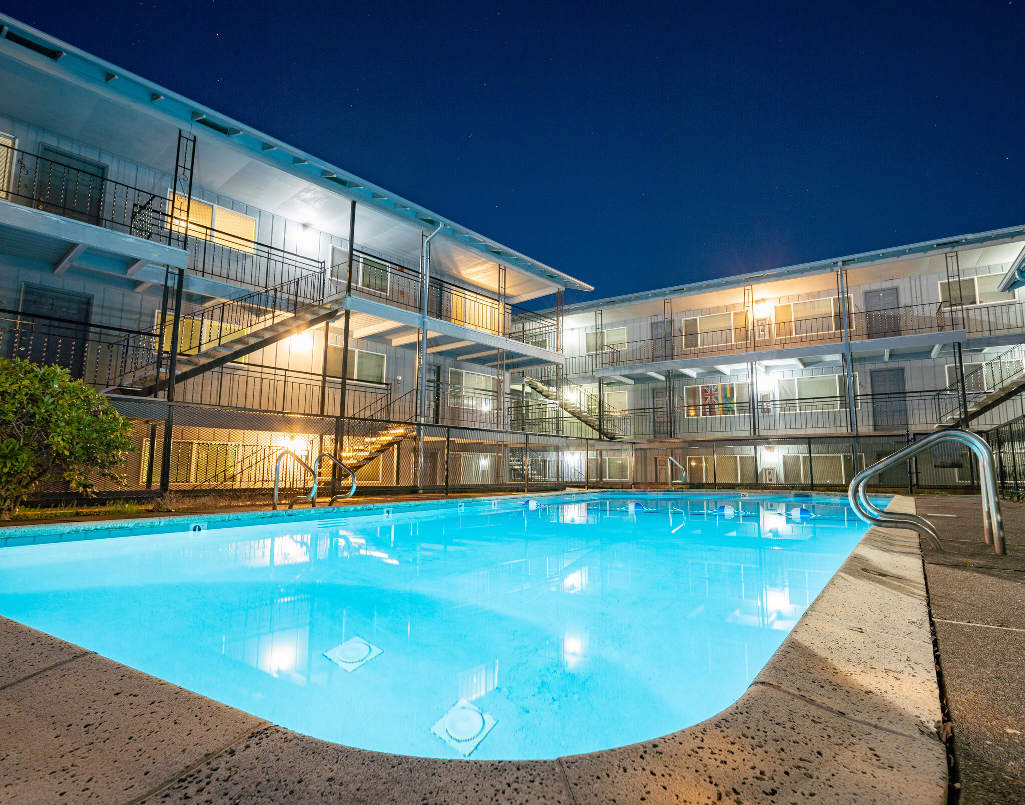 A brightly lit outdoor swimming pool at night is surrounded by a three-story motel called The Vicinity, with glowing windows and balconies. The scene is calm with reflections on the water and a clear, dark sky above.