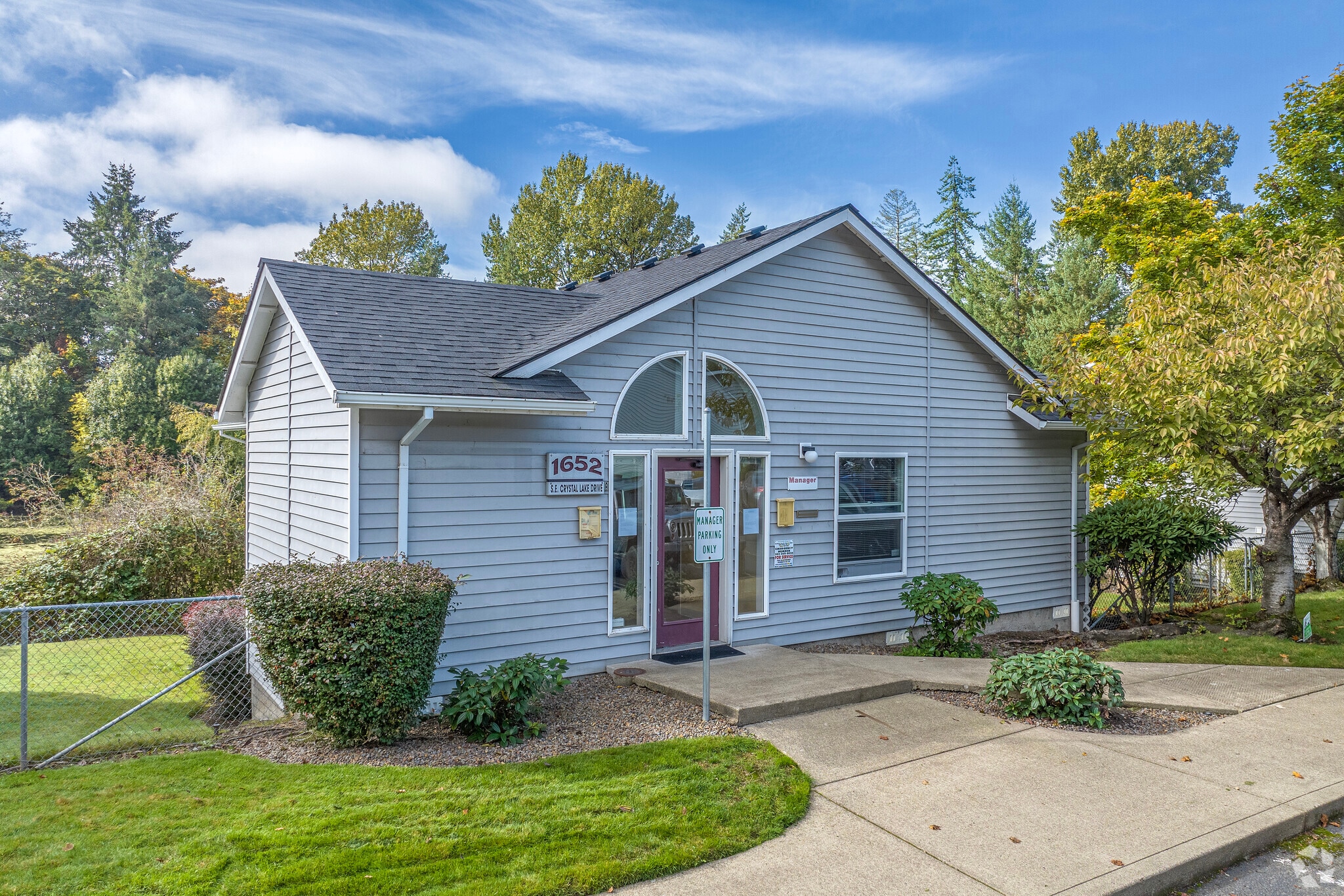 A small gray building with white trim at Crystal Lake Apartments displays the number 1652 above the door. It features a manicured lawn, shrubs, a sidewalk, and is surrounded by trees on a clear, sunny day.
