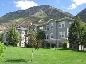 The Harris Condo is a three-story gray apartment building with white trim, surrounded by green lawns and trees, set against a scenic, rocky mountain under a blue sky with scattered clouds.