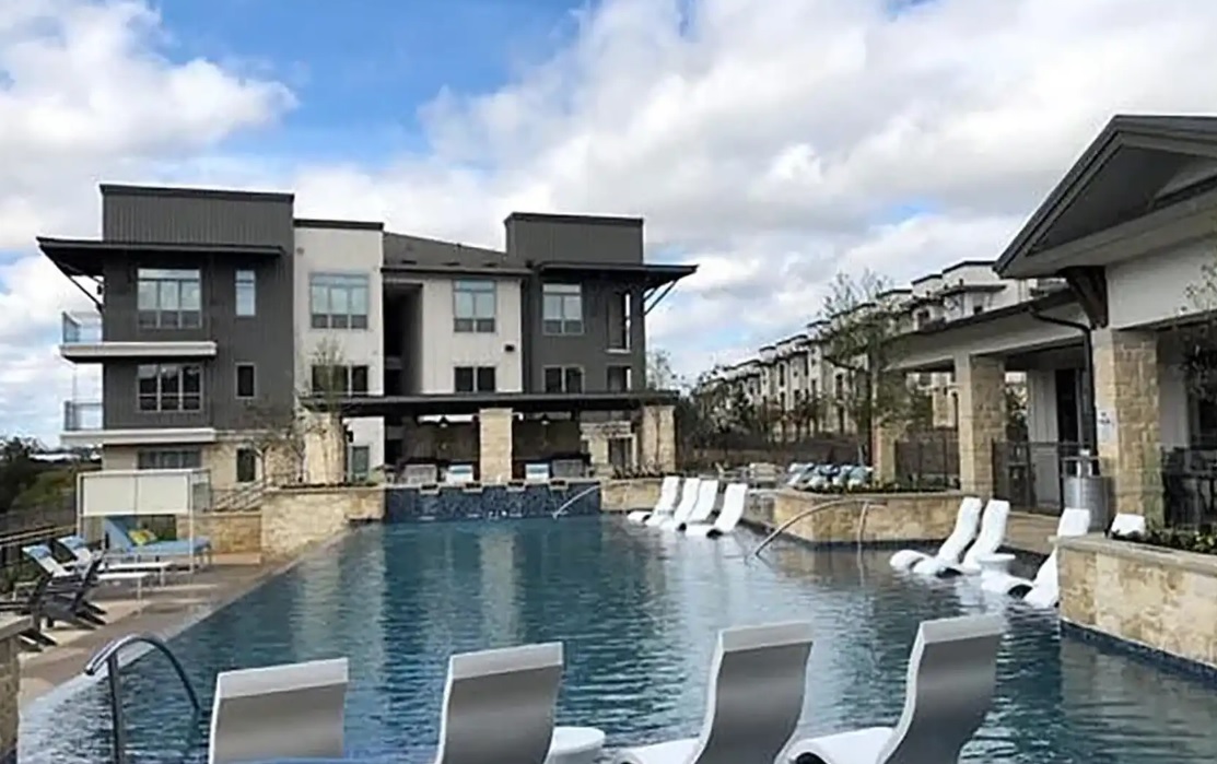 Modern apartment complex, The Kelley at Samuels Avenue Fort Worth, features a large outdoor swimming pool with partially shaded lounge chairs in the water and balconies overlooking the scene under a partly cloudy sky.