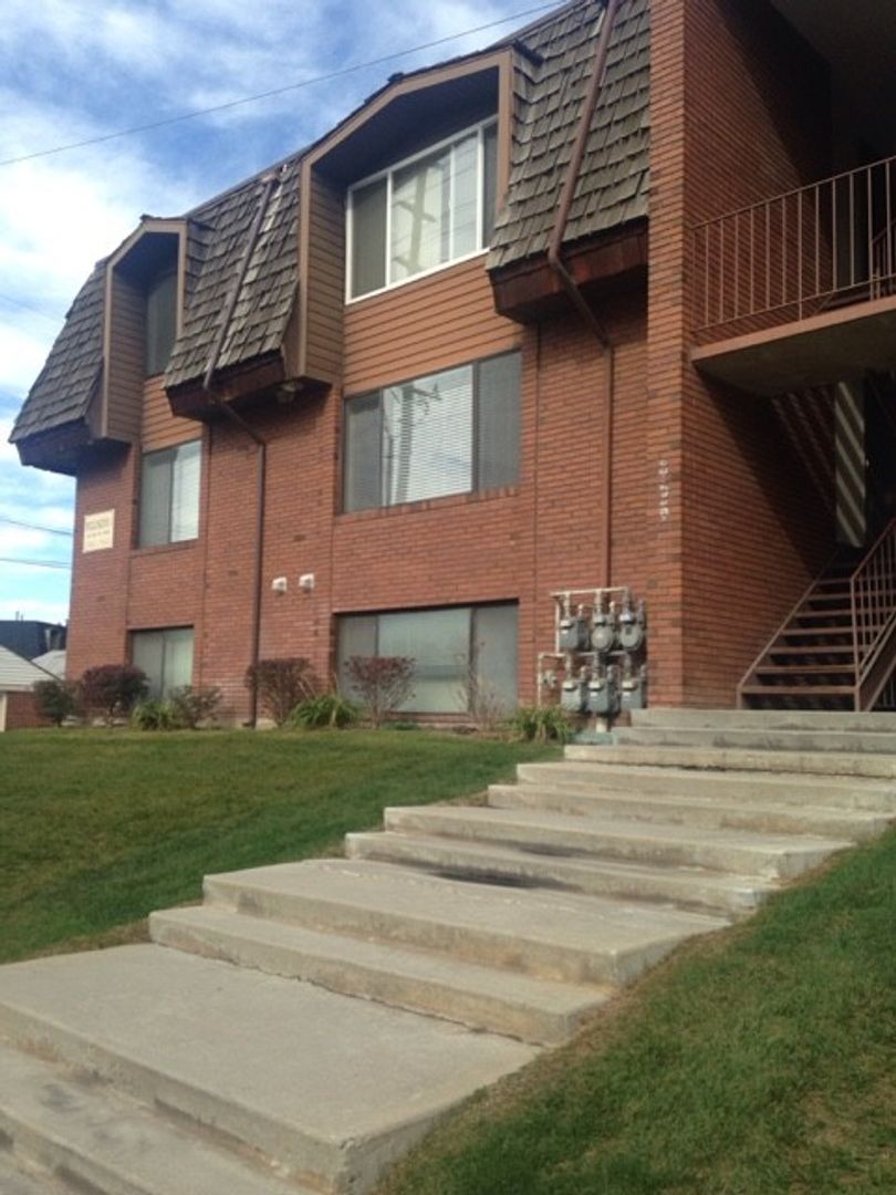 A two-story brick apartment building, known as Wellington Condos by Joseph Thomas, with brown shingle roofs, large windows, exposed gas meters, and a concrete staircase leading up a grassy lawn to the entrance.
