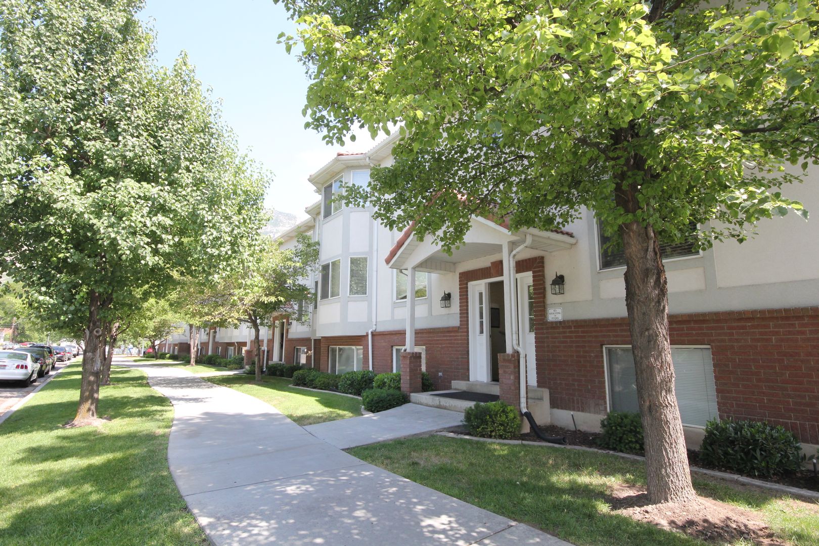 A residential street in Santa Barbara with a sidewalk lined by green trees, featuring modern townhouses with white and red brick exteriors and well-kept lawns on a sunny day, designed by JosephThomas.