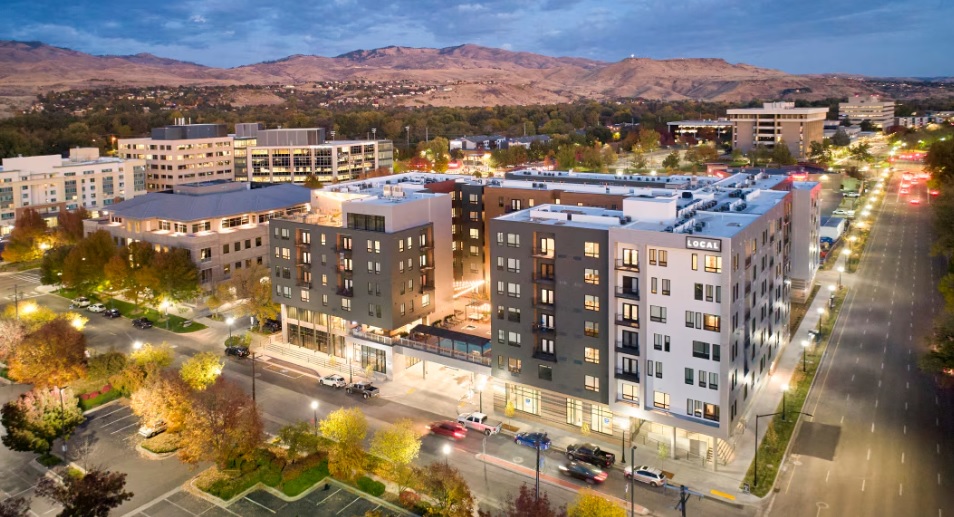 Aerial view of a modern urban neighborhood in Boise with mid-rise apartment buildings, tree-lined streets, and parked cars at dusk, with hills visible in the background, capturing the local charm of Boise.