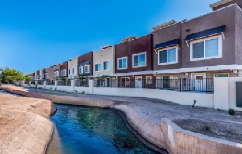 A row of modern two-story townhomes at Lofts on 8th Tempe, with large windows, overlooks a narrow canal under a clear blue sky. The buildings feature varied earth-tone colors and inviting front patios.