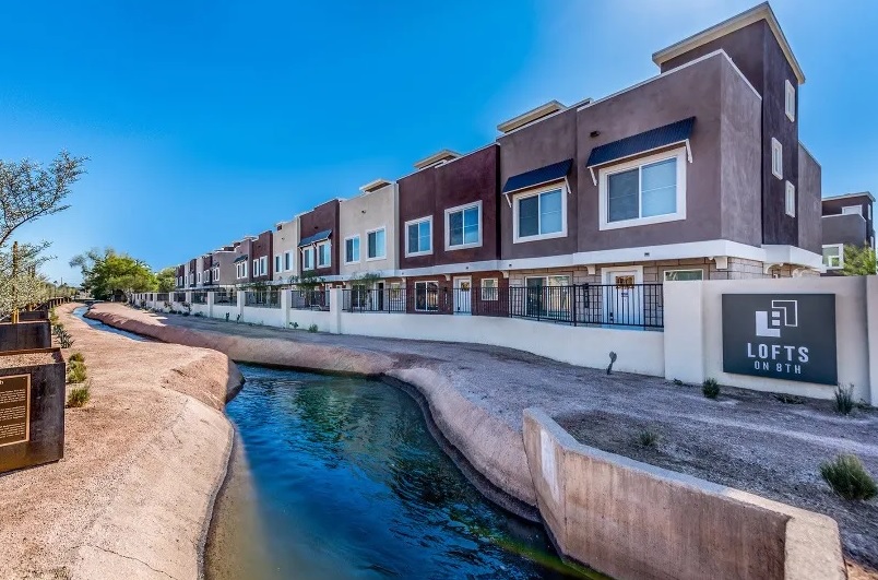 Modern row of brown and beige townhouse-style apartments with large windows, next to a narrow, landscaped canal under a clear blue sky; a sign reads Lofts on 8th Tempe.