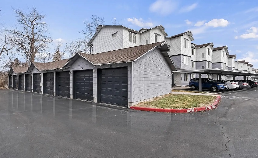 Row of gray single-car garages with dark doors in front of a multi-story apartment complex at Logger Creek at Parkcenter Apartments Boise; covered parking spaces and several parked cars are visible on the right under a blue sky.