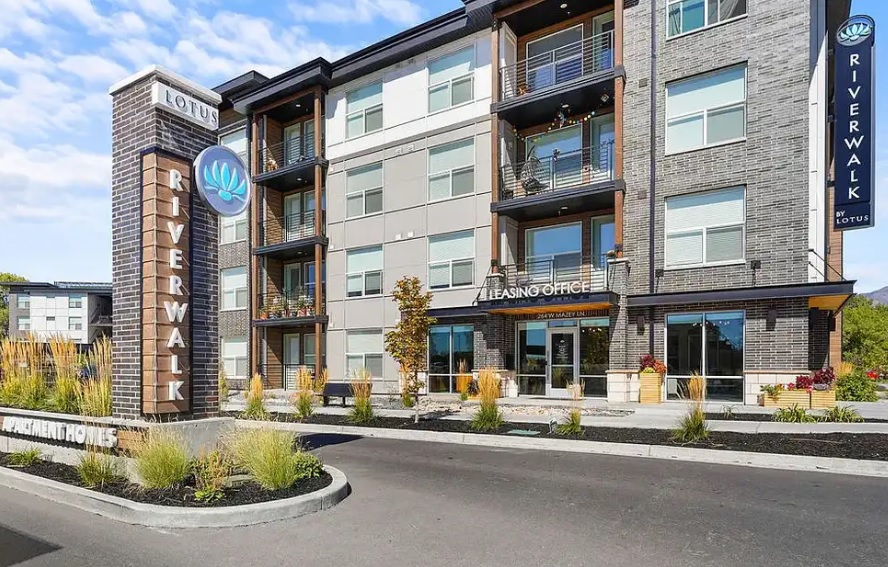 Modern apartment building with a sign reading “RIVERWALK” and a “Leasing Office” entrance. Lotus Riverwalk Ogden features balconies, large windows, and landscaped plants in front beneath a blue sky with scattered clouds.
