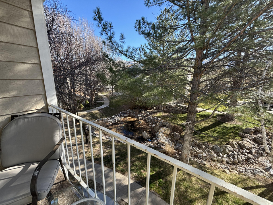 A small balcony at 650 S Main Street with a cushioned chair and glass table overlooks a peaceful, tree-filled park with a winding path and a rocky stream under a clear blue sky.