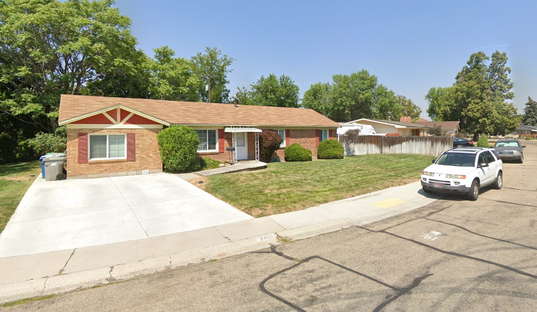 The Malad House Boise is a one-story brick house with a tan roof, front lawn, and small bushes. A white SUV is parked on the street, with trees in the background under a clear blue sky.