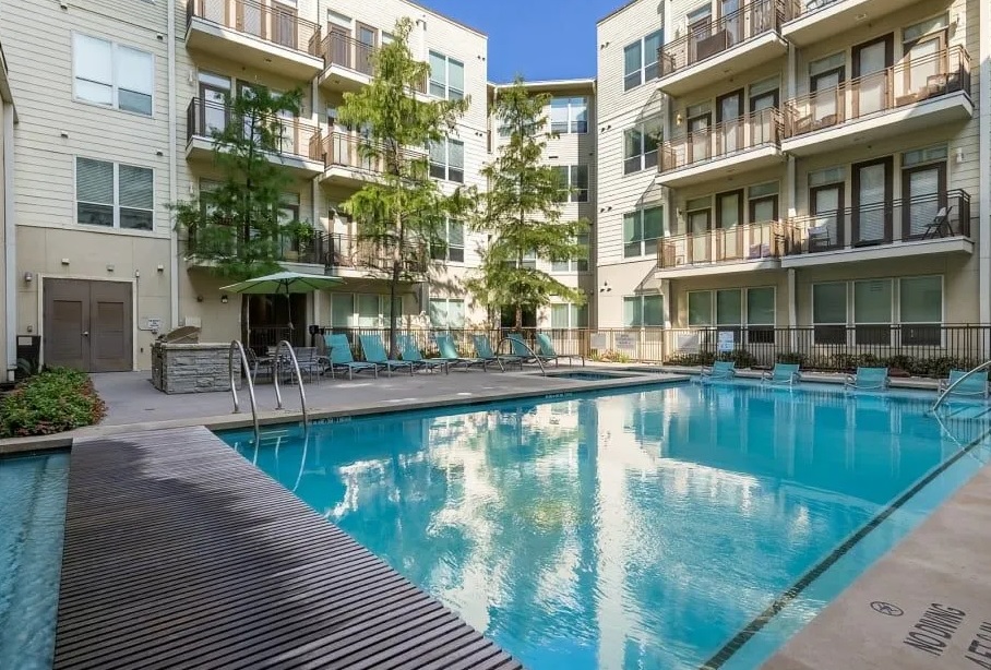 Modern apartment complex courtyard at The Marq on West 7th Fort Worth features a large outdoor swimming pool, lounge chairs, and poolside umbrellas. Surrounding buildings have balconies and large windows, all set beneath clear blue skies.