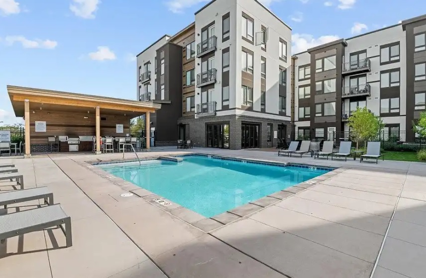 Rectangular outdoor swimming pool surrounded by lounge chairs on a spacious patio at Meritage West Boise, adjacent to modern apartment buildings and a covered seating area, under a blue sky with scattered clouds.