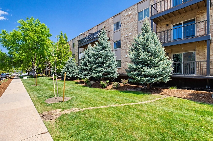 The Mountainaire Boulder apartment building with balconies is surrounded by green lawns, young and mature trees, and a sidewalk under a bright blue sky.