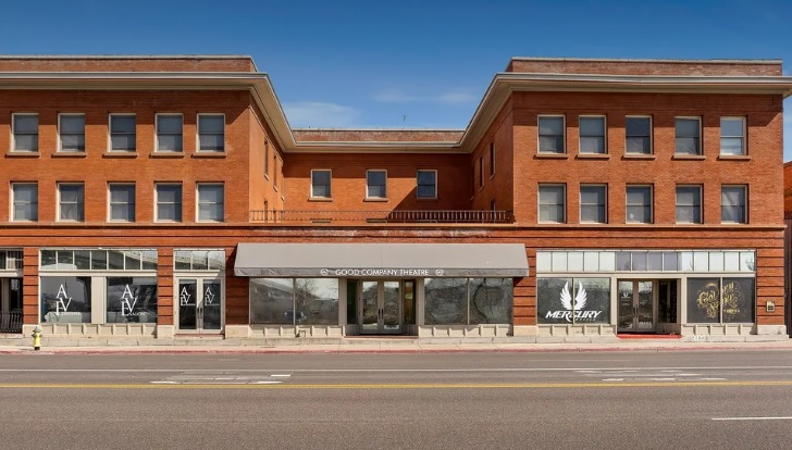 A wide, three-story red brick building with large windows and a central entrance canopy houses the New Brigham Apartments Ogden. The storefronts display signage, including “Mercury” with a white feather logo, beneath the clear blue sky above.