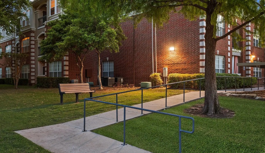 A sidewalk with a metal handrail leads to the brick Olympus 7th Street Station Fort Worth building, passing by a wooden bench and green trees on a grassy lawn. The area is well-lit by exterior wall lights.