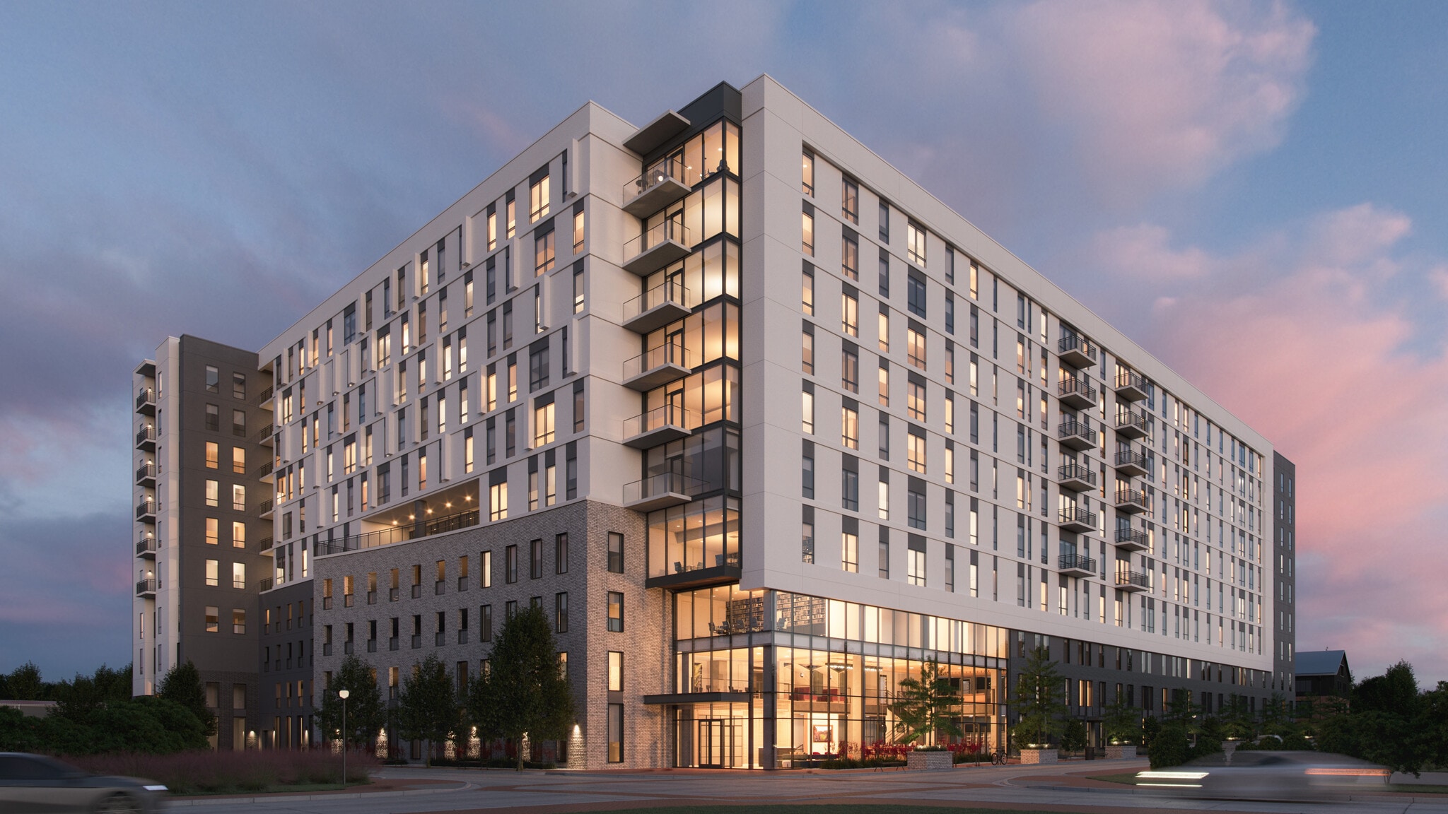 Otto Northgate is a modern mid-rise apartment building with large windows and balconies, illuminated warmly from within at dusk. Cars pass by on the street, and landscaped greenery surrounds the entrance.