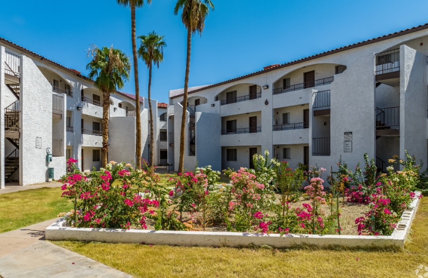 Three-story white Palm Aire Apartments Phoenix buildings surround a central courtyard with blooming pink flowers, palm trees, and a small garden on a sunny day. Balconies and outdoor staircases add charm to the clear blue sky backdrop.