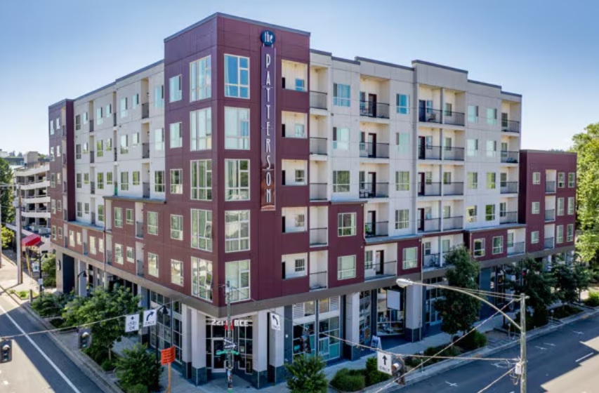 A modern five-story apartment building, The Patterson Social Eugene features red and beige exterior walls, large windows, and balconies. Situated on a street corner with trees and cars nearby, the sign reads The Patterson.