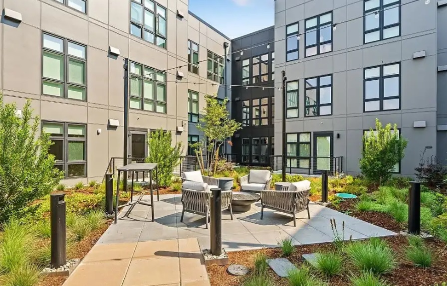 Modern apartment courtyard at Portal Eugene featuring a circular patio with contemporary chairs around a fire pit, surrounded by greenery, mulch, and multi-story beige buildings with large windows.