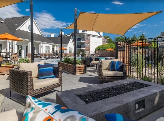 Outdoor lounge area with wicker chairs, cushions, and a rectangular fire pit. Sun shades and orange umbrellas offer shade at The Renaissance at Hobble Creek Boise, with apartment buildings and a swimming pool visible under a blue sky.