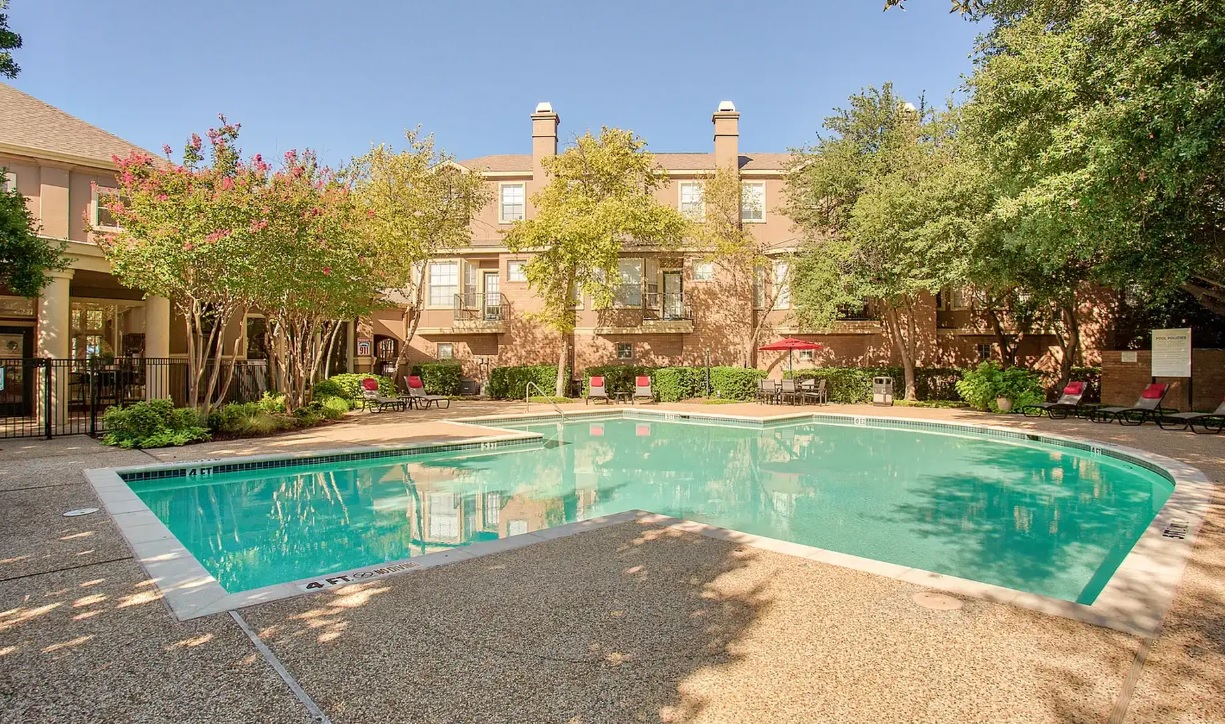 Enjoy the outdoor swimming pool with clear water at The Reserve on Willow Lake Fort Worth, surrounded by lounge chairs, trees, and a multi-story brick apartment building under a clear blue sky.