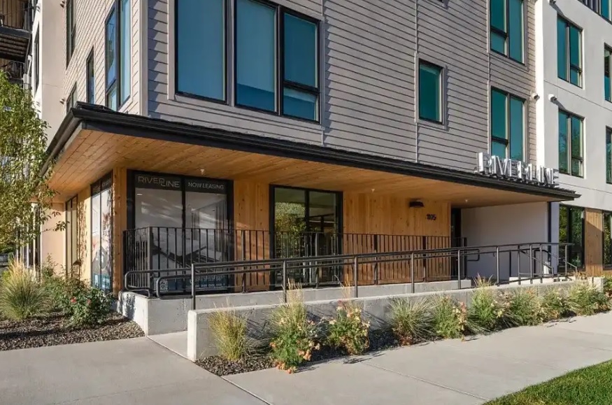 Modern apartment building with large windows, a wooden facade, and a ramp leading to the entrance. The sign above reads RIVERINE, highlighting its connection to Riverline Boise. Landscaping features grass and shrubs along the sidewalk.
