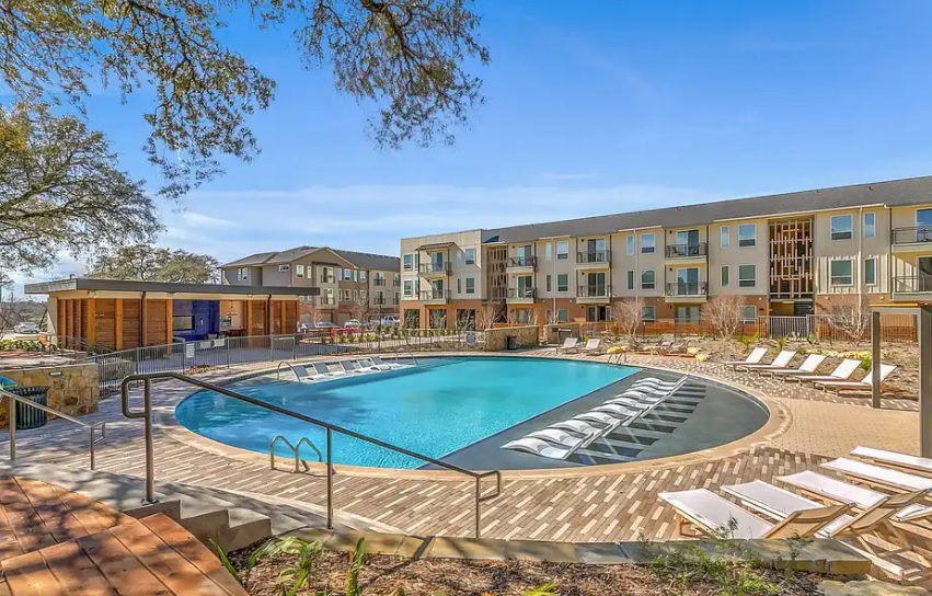 Experience Scenic at River East Fort Worth—a modern apartment complex featuring a large outdoor swimming pool, a wooden deck with lounge chairs, and tree-lined views. Three-story buildings with balconies complete the inviting scene under a clear blue sky.