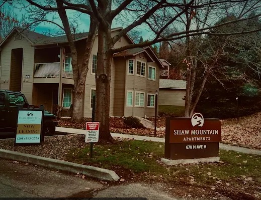 A tan apartment building with balconies is surrounded by trees. A sign in front reads Shaw Mountain Apartments Boise, 670 N Ave H. Another sign nearby says Now Leasing with a phone number. Fallen leaves cover the ground.