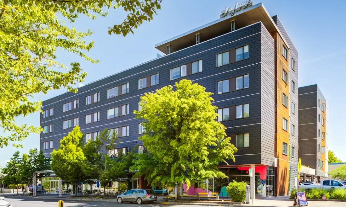 A modern, multi-story hotel building with large windows and a dark exterior, Yugo Eugene Skybox stands tall, surrounded by green trees and parked cars on a sunny day.