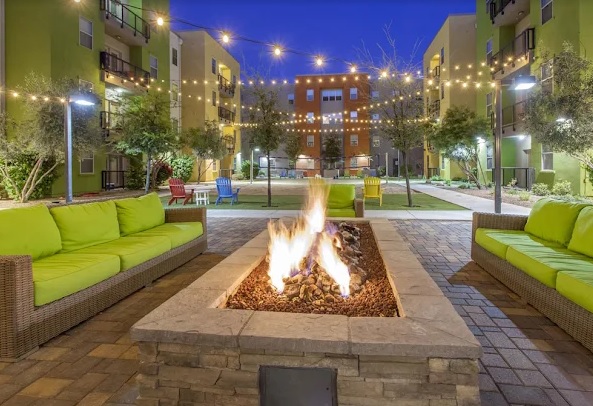A modern courtyard at Sol Tempe Apartments at dusk features green cushioned sofas around a rectangular stone fire pit, string lights overhead, and colorful chairs in the background—perfect for relaxing evenings in Tempe.