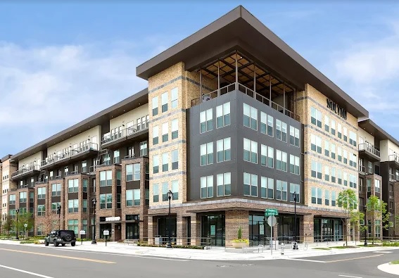 Modern multi-story South 400 Apartments Fort Worth building on a city corner with large windows, balconies, and a mix of brick and dark exterior panels. Trees and parked cars line the street under a clear sky.