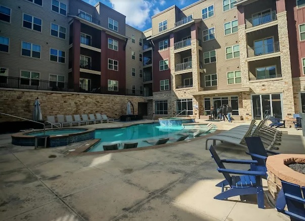 Outdoor pool area at Stadium Apartments Fort Collins with lounge chairs, a hot tub, and seating around a fire pit. Multi-story buildings with balconies and windows surround the space while some people relax poolside.