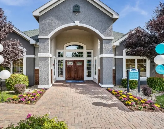 A gray building with white trim, arched entrance, large double wooden doors, and a brick walkway surrounded by colorful flower beds, bushes, and balloons at The Highlands Reno.
