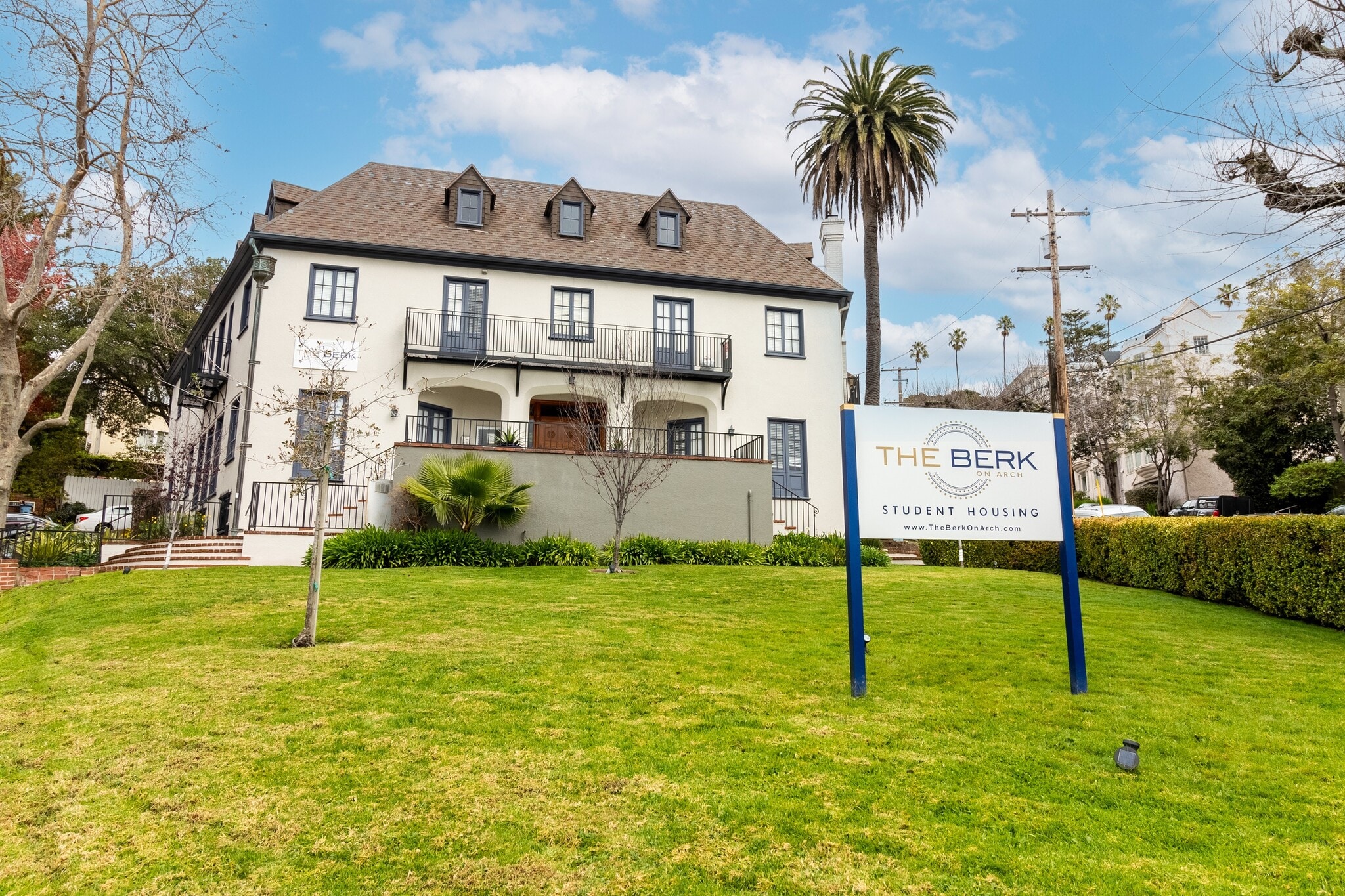 A large, three-story house with dormer windows and a manicured lawn sits behind a sign reading The Berk Student Housing on a sunny day with palm trees and blue sky in the background.