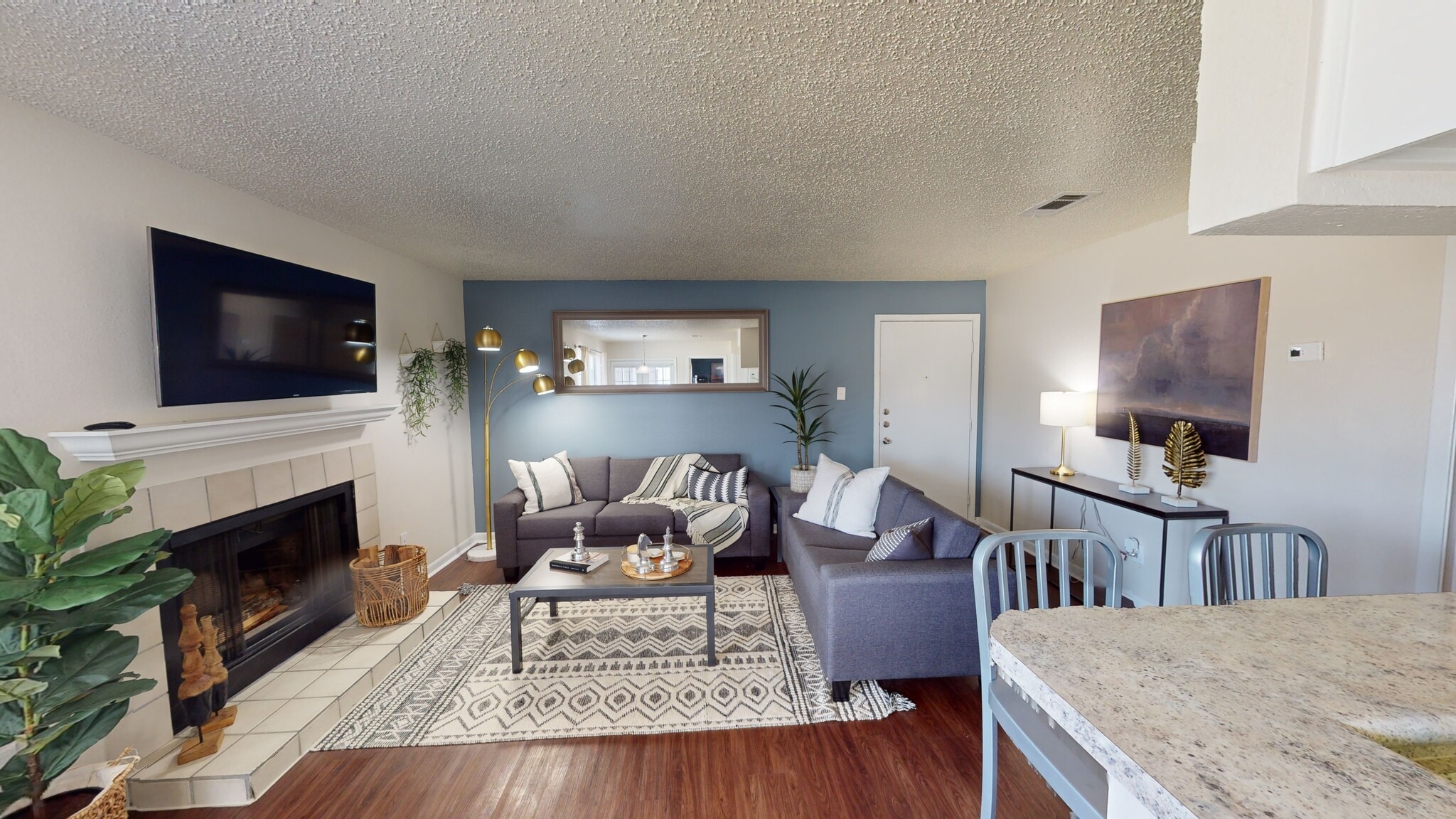A modern living room in Landing College Station features a gray sectional sofa, patterned rug, coffee table, fireplace with TV above, wall mirror, and indoor plants. Wooden flooring and a light blue accent wall create a stylish student housing vibe.