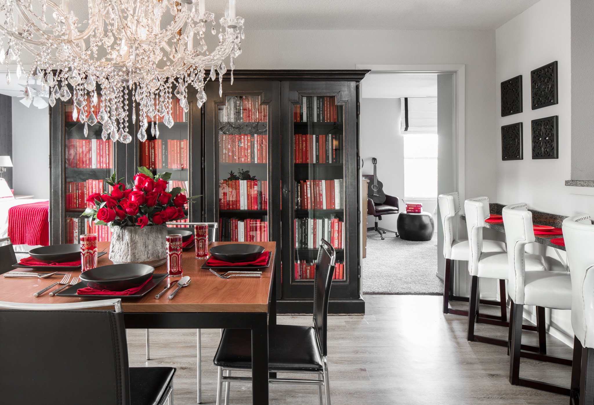 Modern dining room with a wooden table set for four, black and red dishware, a crystal chandelier, black bookshelves filled with red and white books, and a bright adjoining room with a guitar visible.