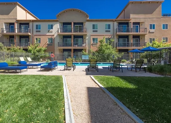 The Social at Stadium Walk Fort Collins features a modern three-story apartment building overlooking an outdoor swimming pool with lounge chairs, umbrellas, and a grassy area under a clear blue sky.