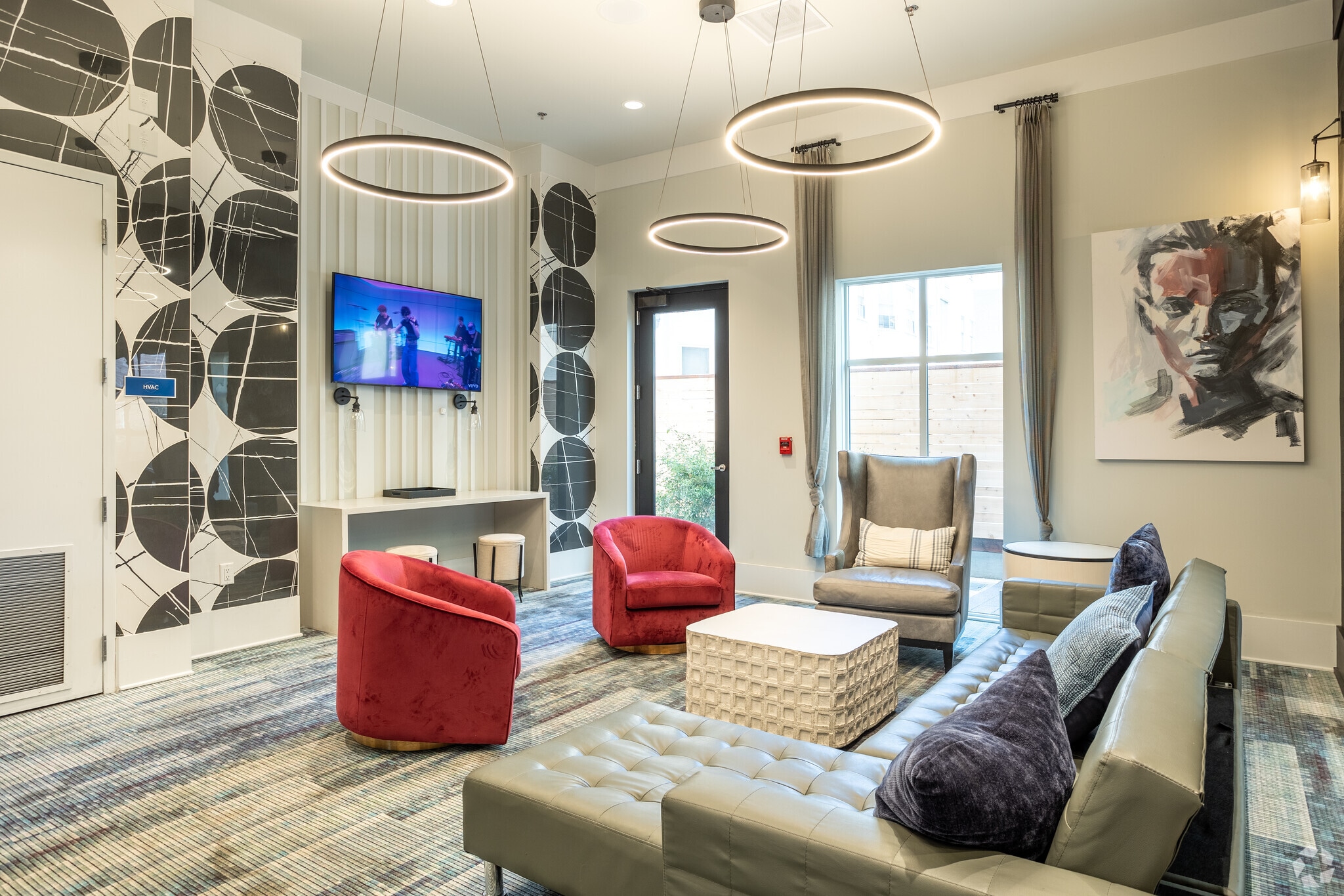 Modern lounge at The Standard at Berkeley with a gray sectional sofa, two red armchairs, a high-back gray chair, wall-mounted TV, abstract art, circular hanging lights, and large windows letting in natural light.