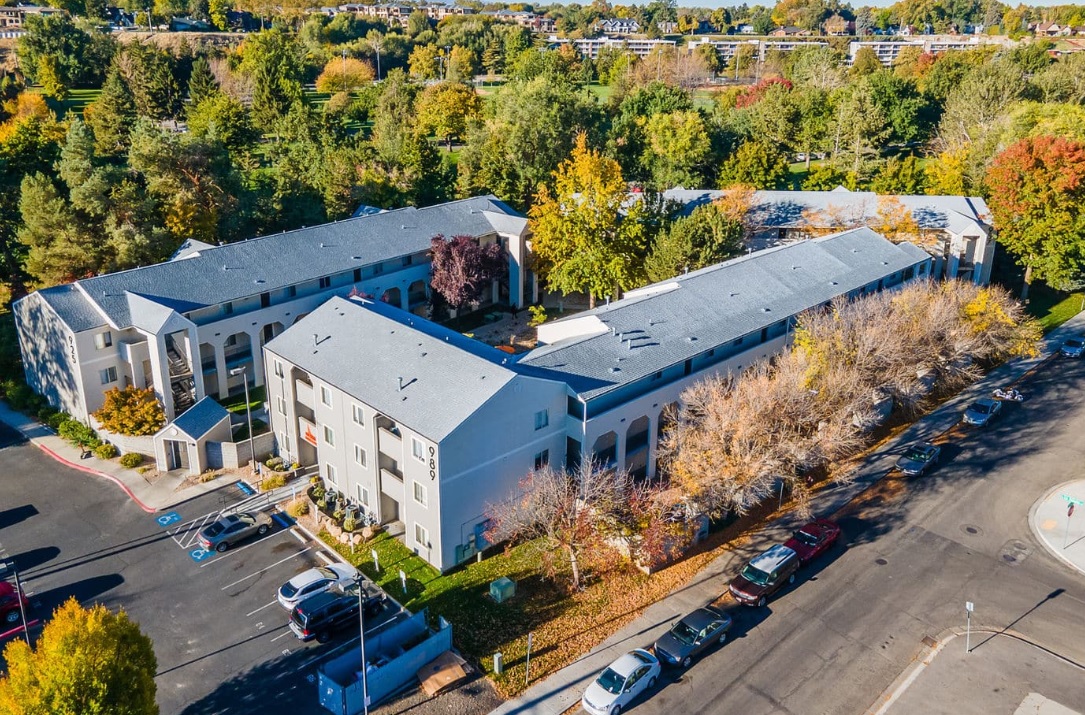 Aerial view of The 208 Boise, a large U-shaped apartment complex surrounded by autumn trees. Cars fill the street and a lot with marked accessible spaces. Nearby streets and buildings complete the scene.