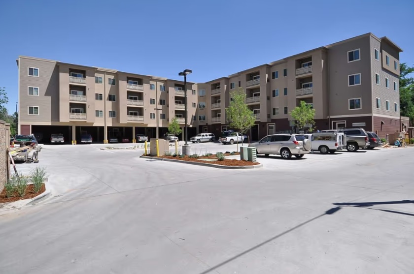 Flats at the Oval Fort Collins is a modern, four-story apartment building with balconies and a parking lot in front, featuring several parked cars and small landscaped areas, all set on a clear, sunny day.