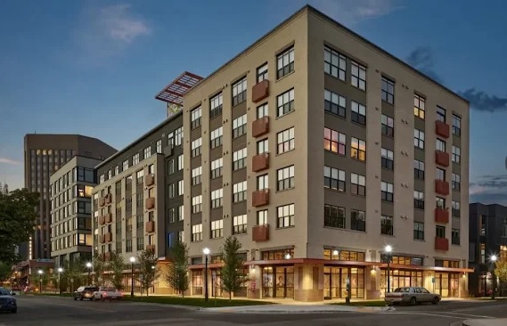 A modern, multi-story apartment building with large windows is shown at dusk. Streetlights are on, and a few cars are parked along the tree-lined sidewalk in front of The Lucy Boise.