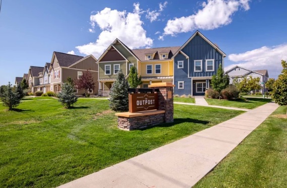 A row of colorful modern houses lines a quiet suburban street with green lawns and young trees. A stone sign in front reads The Outpost Fort Collins. The sky is blue with scattered white clouds.
